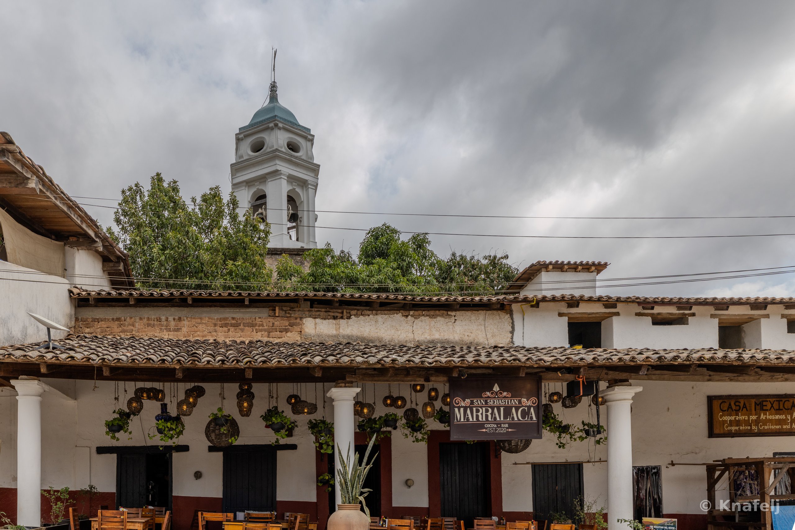 A colonial-style building with white columns, tiled roof, and hanging lanterns displays a sign reading Marralaca, while a white church bell tower with a blue dome rises behind it under an overcast sky.