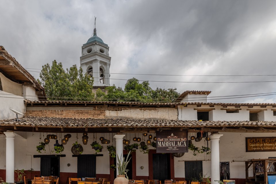 A colonial-style building with white columns, tiled roof, and hanging lanterns displays a sign reading Marralaca, while a white church bell tower with a blue dome rises behind it under an overcast sky.