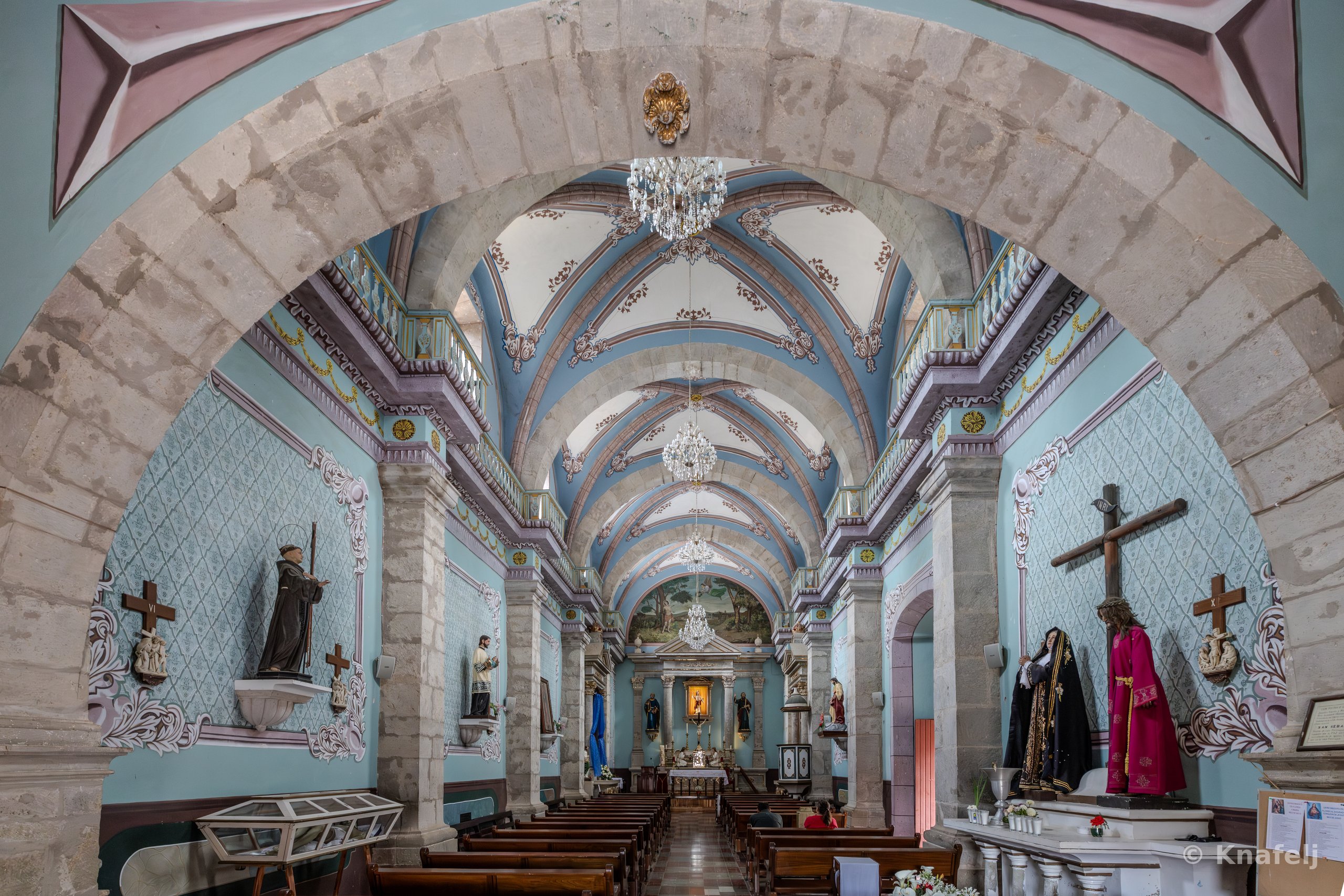Interior of a colonial church viewed through a large stone arch, showing a long nave with blue vaulted ceilings, crystal chandeliers, religious statues, a crucifix on the right wall, wooden pews, and a distant altar.