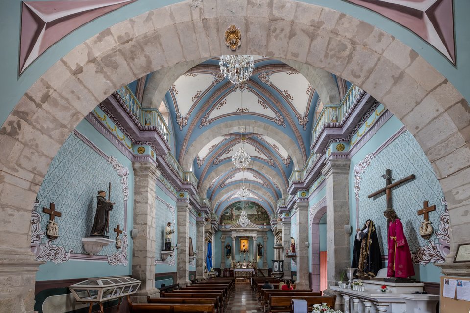 Interior of a colonial church viewed through a large stone arch, showing a long nave with blue vaulted ceilings, crystal chandeliers, religious statues, a crucifix on the right wall, wooden pews, and a distant altar.