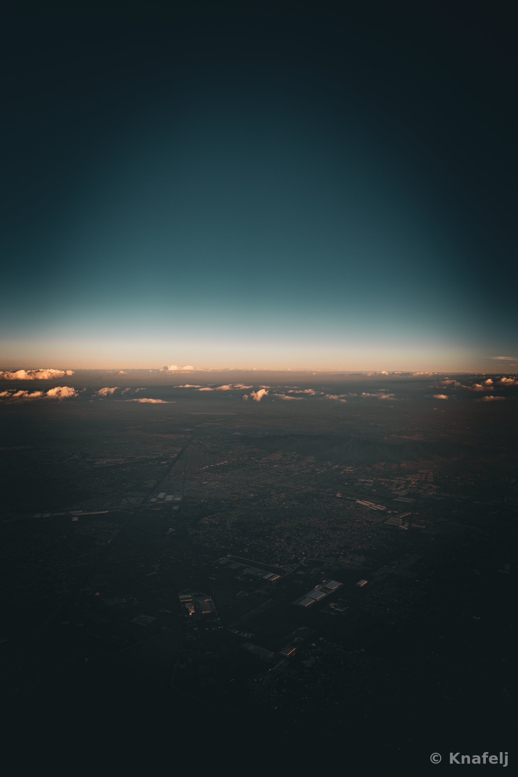 Aerial photograph taken from an airplane showing a vast city landscape below, a horizon line lit with orange glow, low clouds, and a gradient sky transitioning from teal to dark navy.