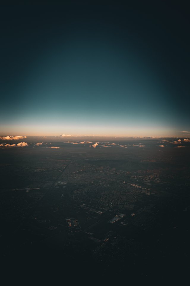 Aerial photograph taken from an airplane showing a vast city landscape below, a horizon line lit with orange glow, low clouds, and a gradient sky transitioning from teal to dark navy.