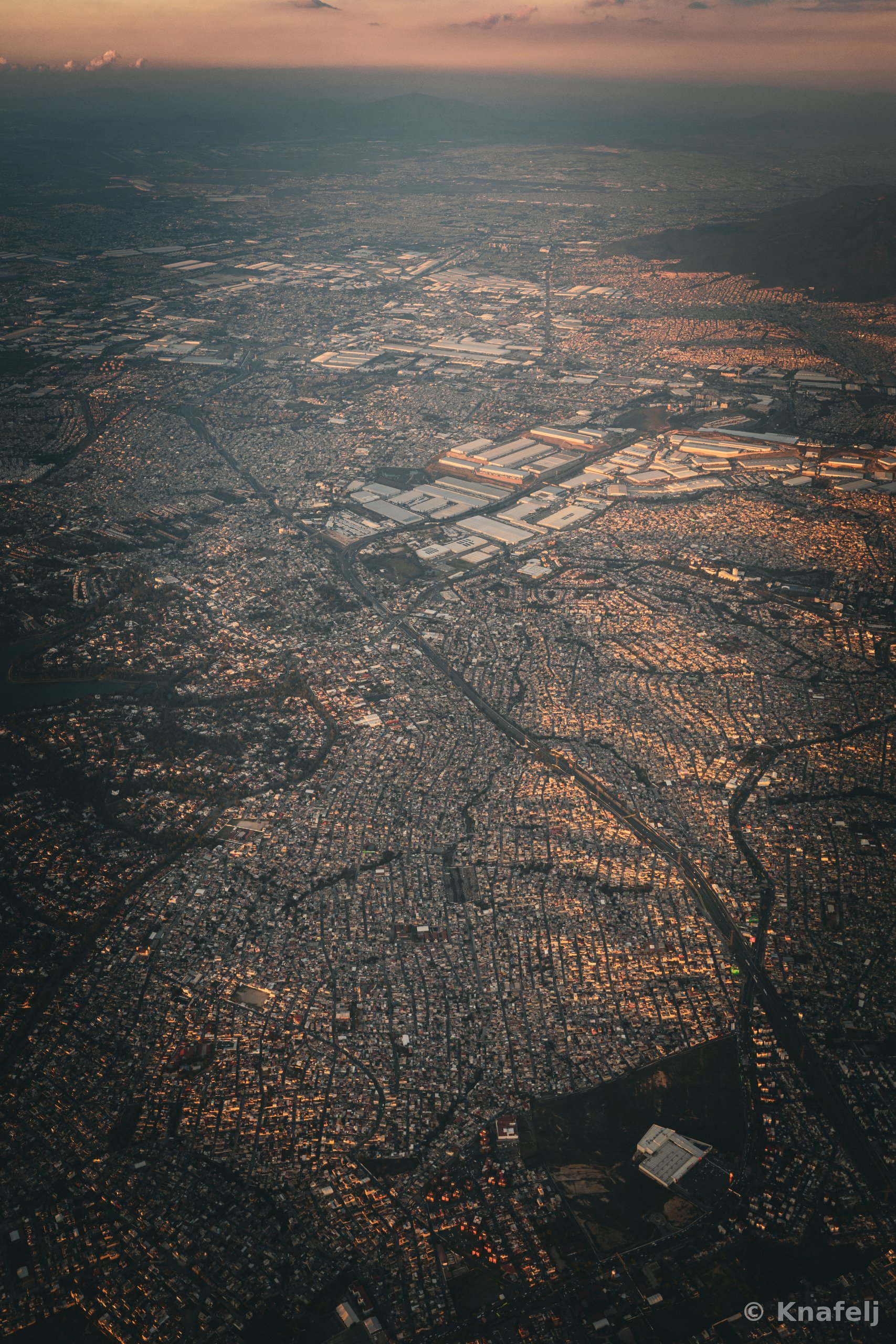 High-altitude aerial photograph of a sprawling city seen from an airplane, with dense residential grids, large industrial warehouse rooftops, roads and highways cutting across the landscape, and hazy mountains along the horizon under a soft twilight sky.