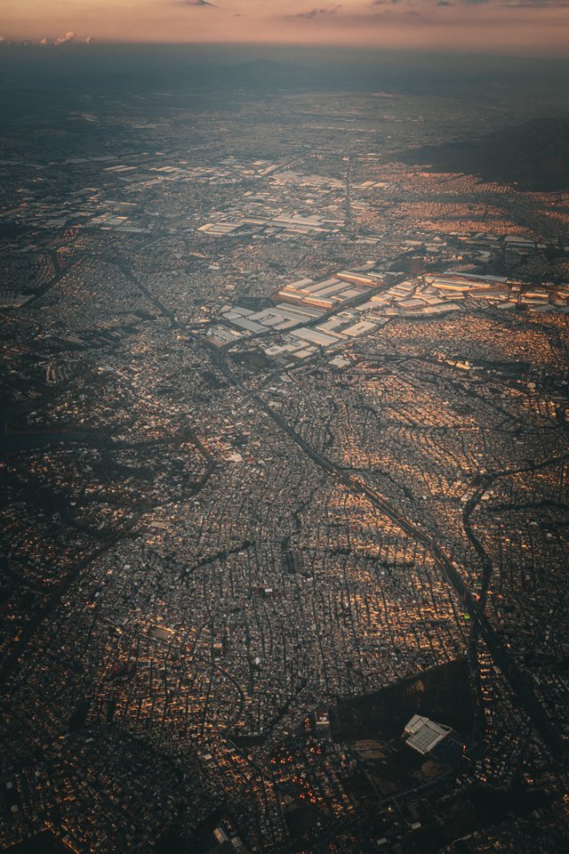High-altitude aerial photograph of a sprawling city seen from an airplane, with dense residential grids, large industrial warehouse rooftops, roads and highways cutting across the landscape, and hazy mountains along the horizon under a soft twilight sky.