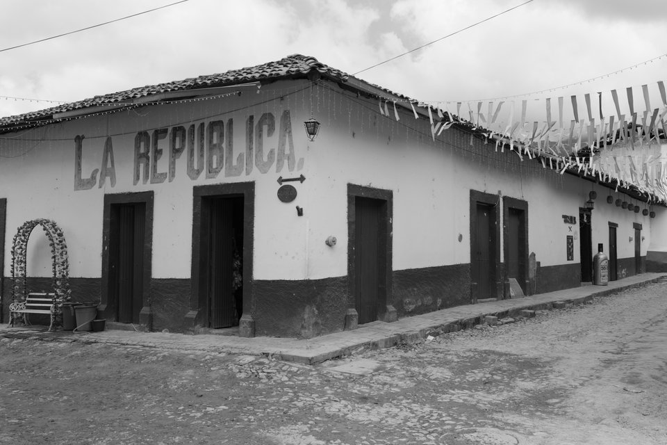 Black and white photo of a single-story corner building with whitewashed walls reading 'LA REPUBLICA' in large painted letters, a tiled roof, dark wooden doors, paper streamers strung overhead, and a cobblestone street in the foreground.
