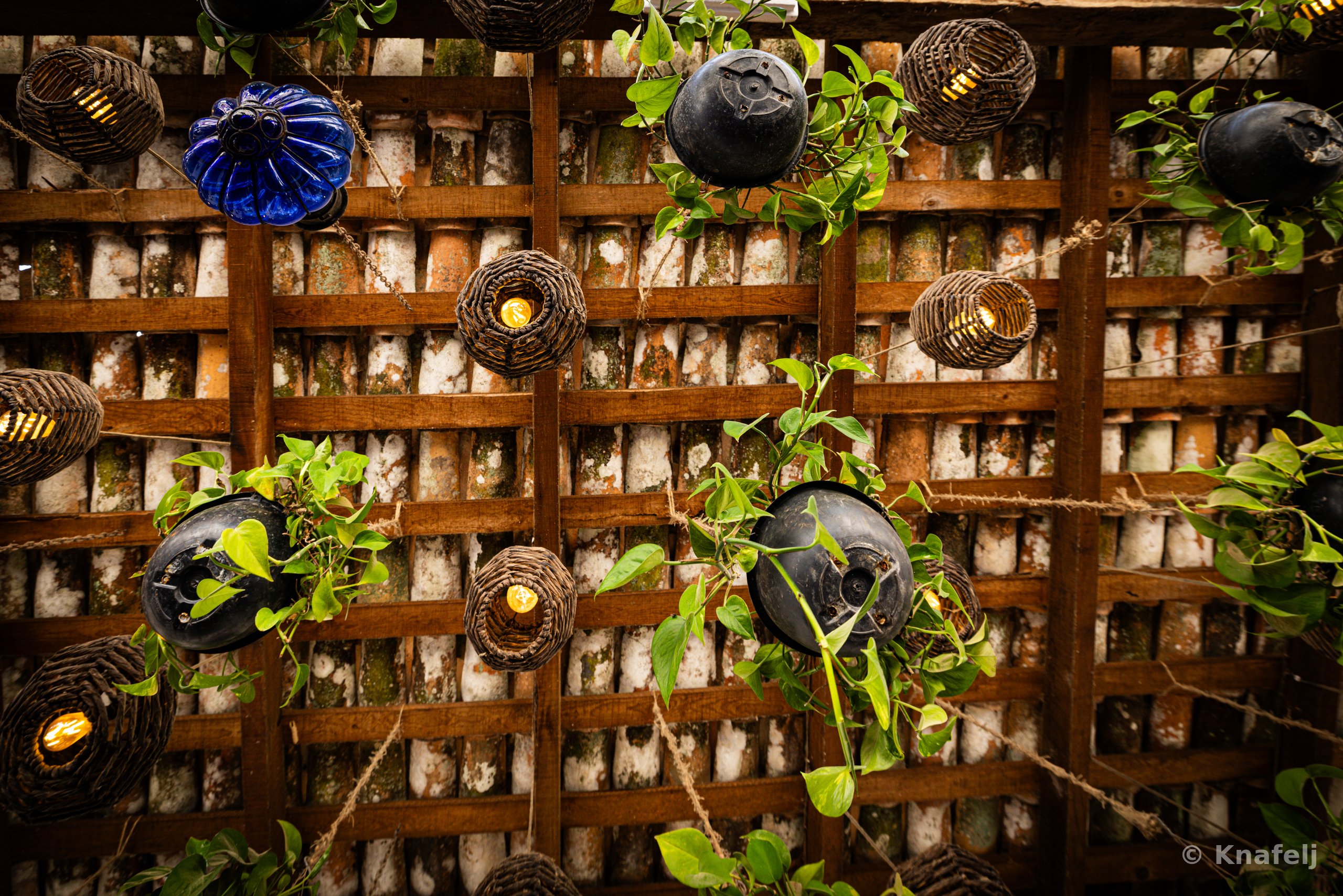 A wooden lattice over a brick wall decorated with various hanging lanterns including woven rattan globes, a blue glass ornament, and dark ceramic orbs, with green pothos vines trailing throughout and small lit bulbs glowing.