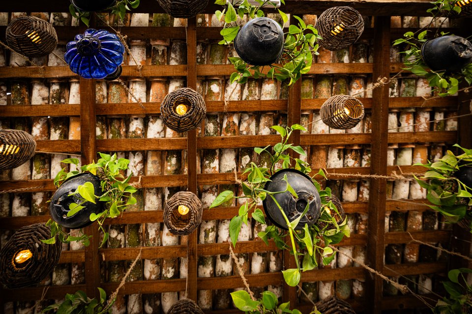 A wooden lattice over a brick wall decorated with various hanging lanterns including woven rattan globes, a blue glass ornament, and dark ceramic orbs, with green pothos vines trailing throughout and small lit bulbs glowing.
