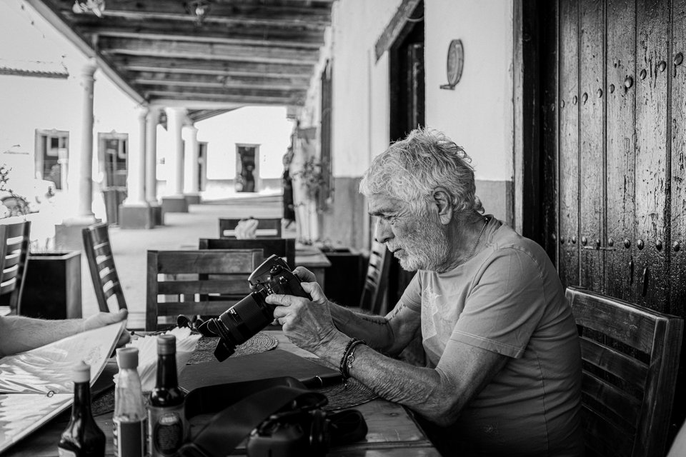Black and white photo of an elderly man with gray hair and beard seated at a table on a covered porch, holding and looking at a DSLR camera. Bottles and a notebook rest on the table, with columns and chairs along a colonial walkway in the background.
