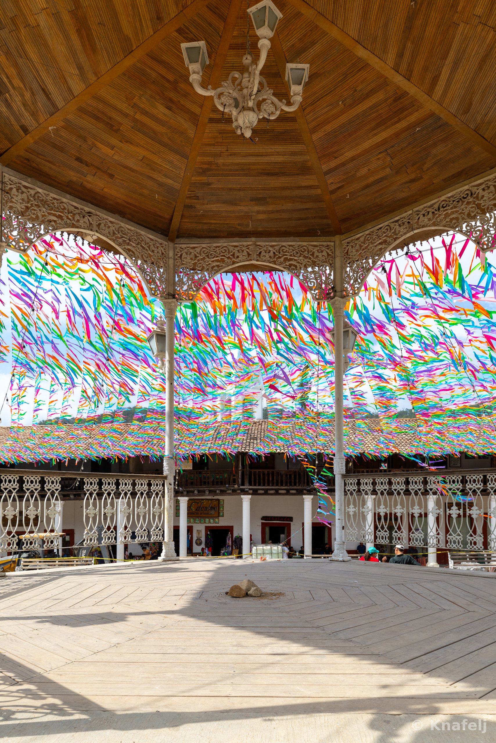 Interior view of a large wooden gazebo or pavilion with a peaked timber ceiling and white chandelier, surrounded by ornate white columns and railings. Colorful ribbons in many colors are strung across the open sky beyond, above a tiled rooftop and a sunlit plank floor.