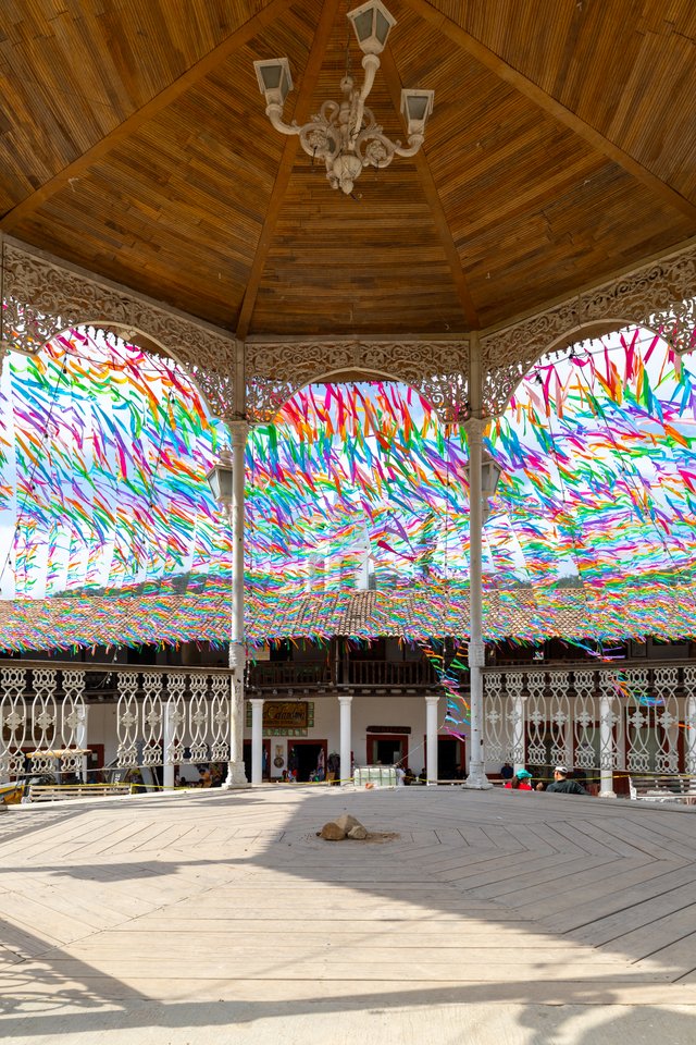 Interior view of a large wooden gazebo or pavilion with a peaked timber ceiling and white chandelier, surrounded by ornate white columns and railings. Colorful ribbons in many colors are strung across the open sky beyond, above a tiled rooftop and a sunlit plank floor.