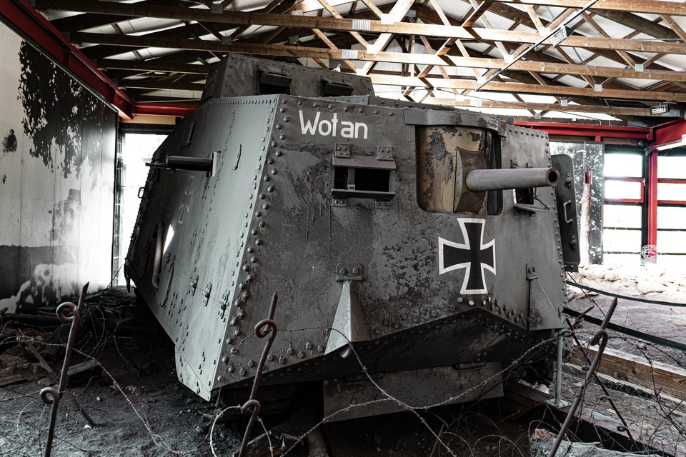 A large grey World War I German tank with the name 'Wotan' and an Iron Cross painted on its side, displayed inside a building with an exposed wooden roof and tangled barbed wire in the foreground.