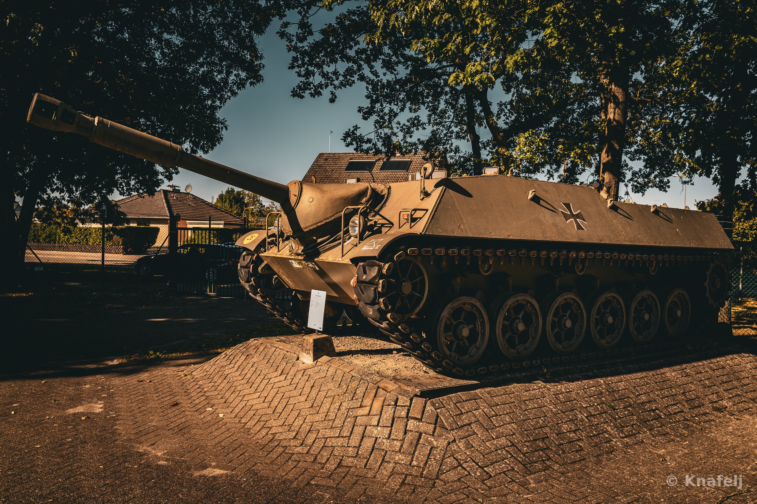 A military tank with a long gun barrel mounted on a brick paved platform, surrounded by trees and houses in the background under a clear sky.