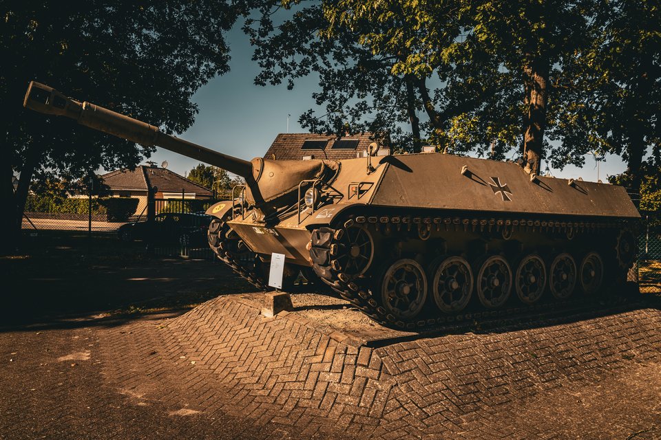 A military tank with a long gun barrel mounted on a brick paved platform, surrounded by trees and houses in the background under a clear sky.