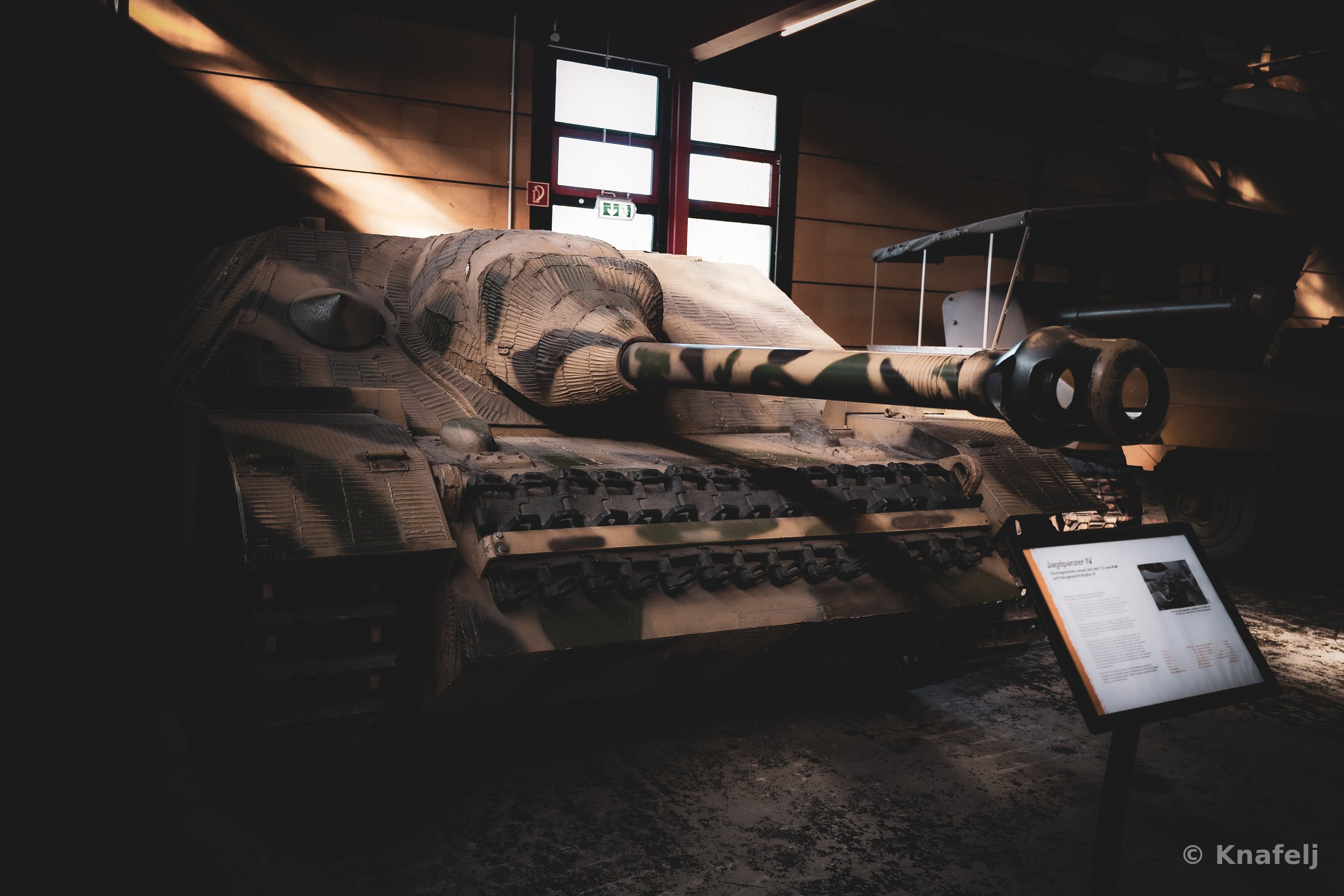 A camouflage-painted military tank with a long gun barrel displayed indoors in a museum, with an informational placard in the foreground and light coming through windows in the background.