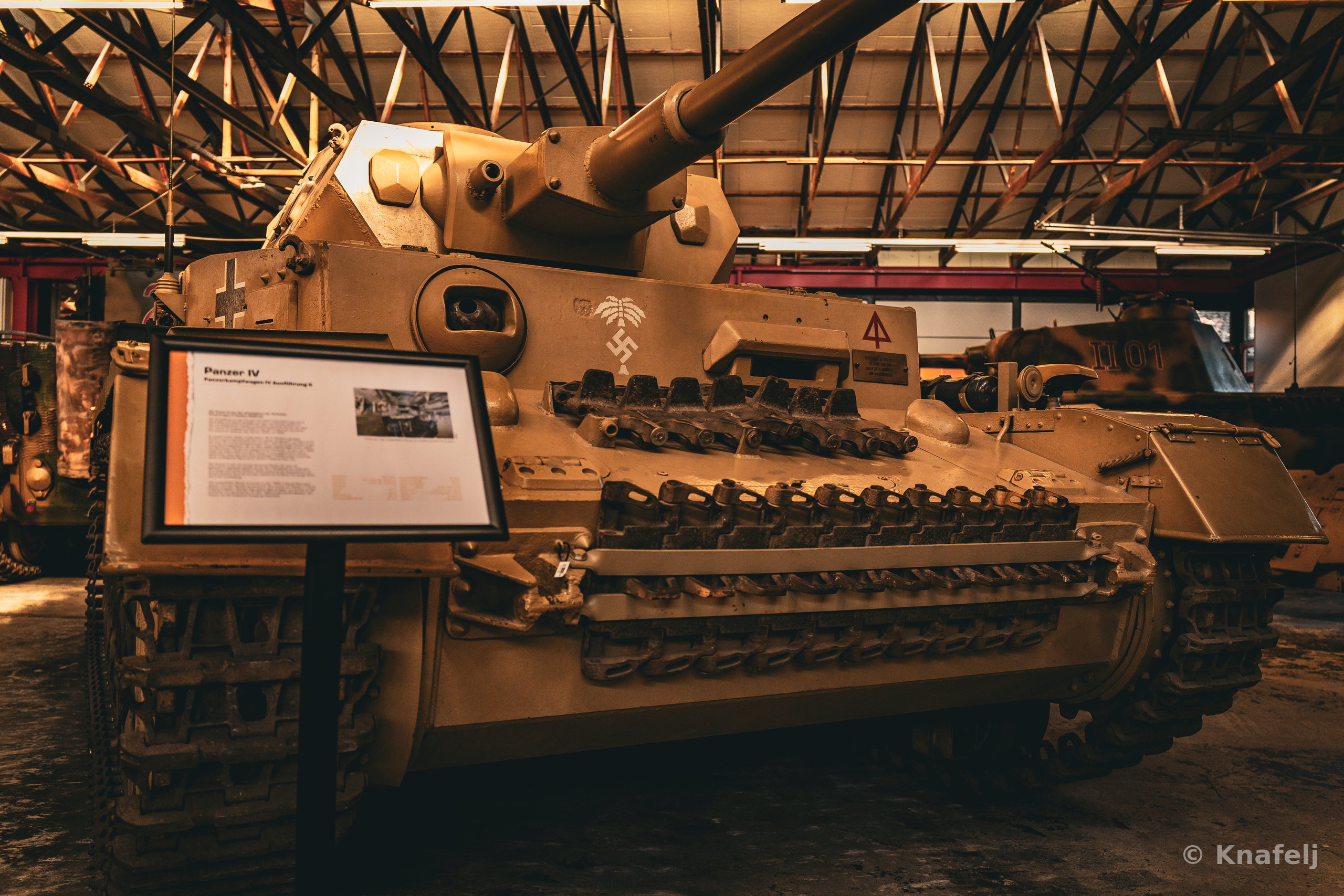 A tan World War II Panzer IV tank with a long gun barrel displayed indoors in a museum, with an information sign in front and steel roof trusses overhead.