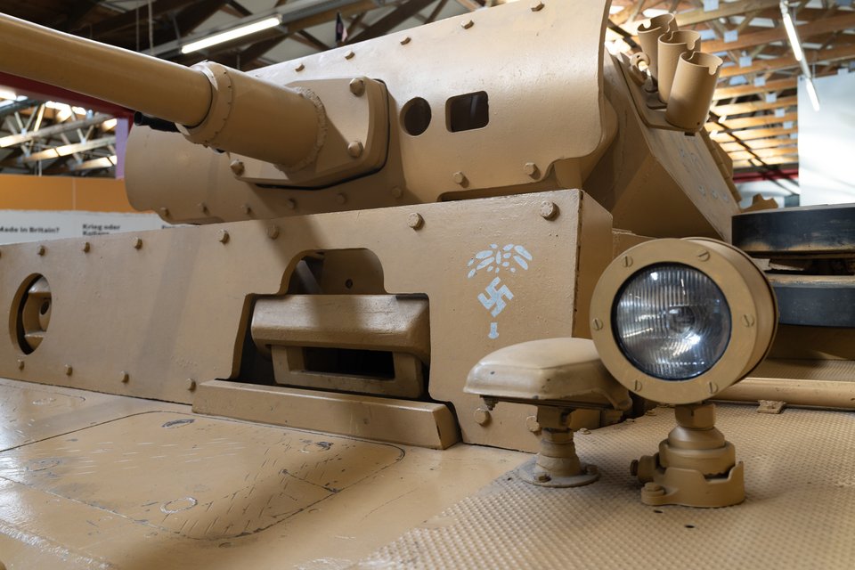 A tan-colored armored military vehicle with a long gun barrel and a round headlight, marked with a faded white emblem, displayed indoors under a wooden-trussed roof.