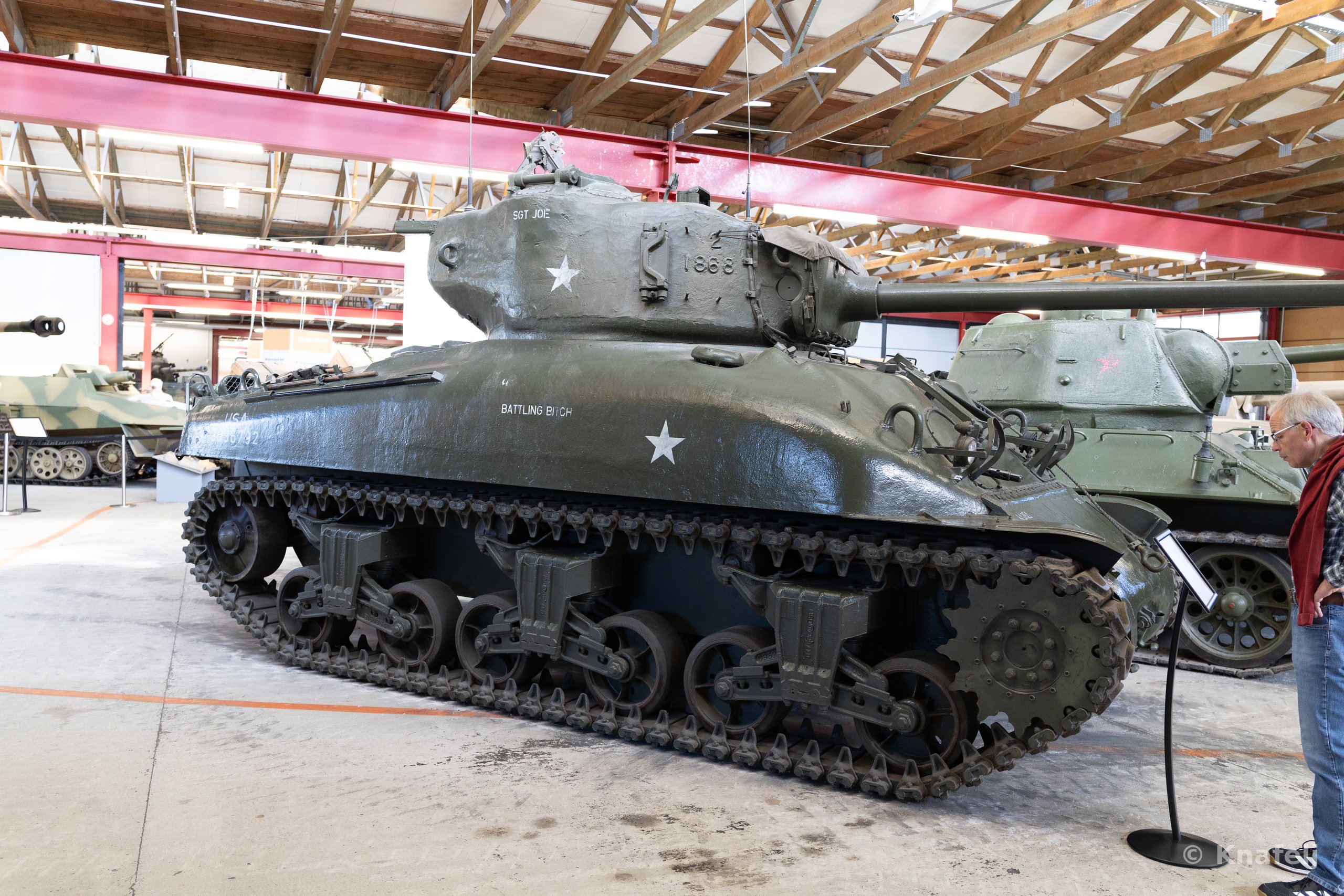 An olive-green Sherman tank with white star markings displayed indoors in a museum hall with wooden roof beams. A Soviet tank stands behind it, and a man in dark clothing reads an information panel to the right.