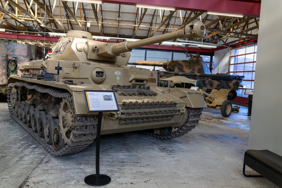 A sand-colored German Panzer III tank displayed indoors in a museum hangar, with tracks and a raised main gun, surrounded by other military vehicles and an information sign.