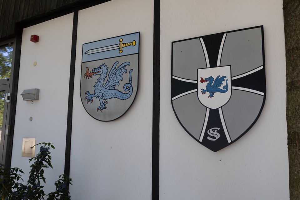 Two coats of arms displayed on a white building wall with black trim. The left shield shows a blue dragon below a sword on a light blue band; the right shield features a blue dragon on a white inset against gray and dark panels divided by a cross.