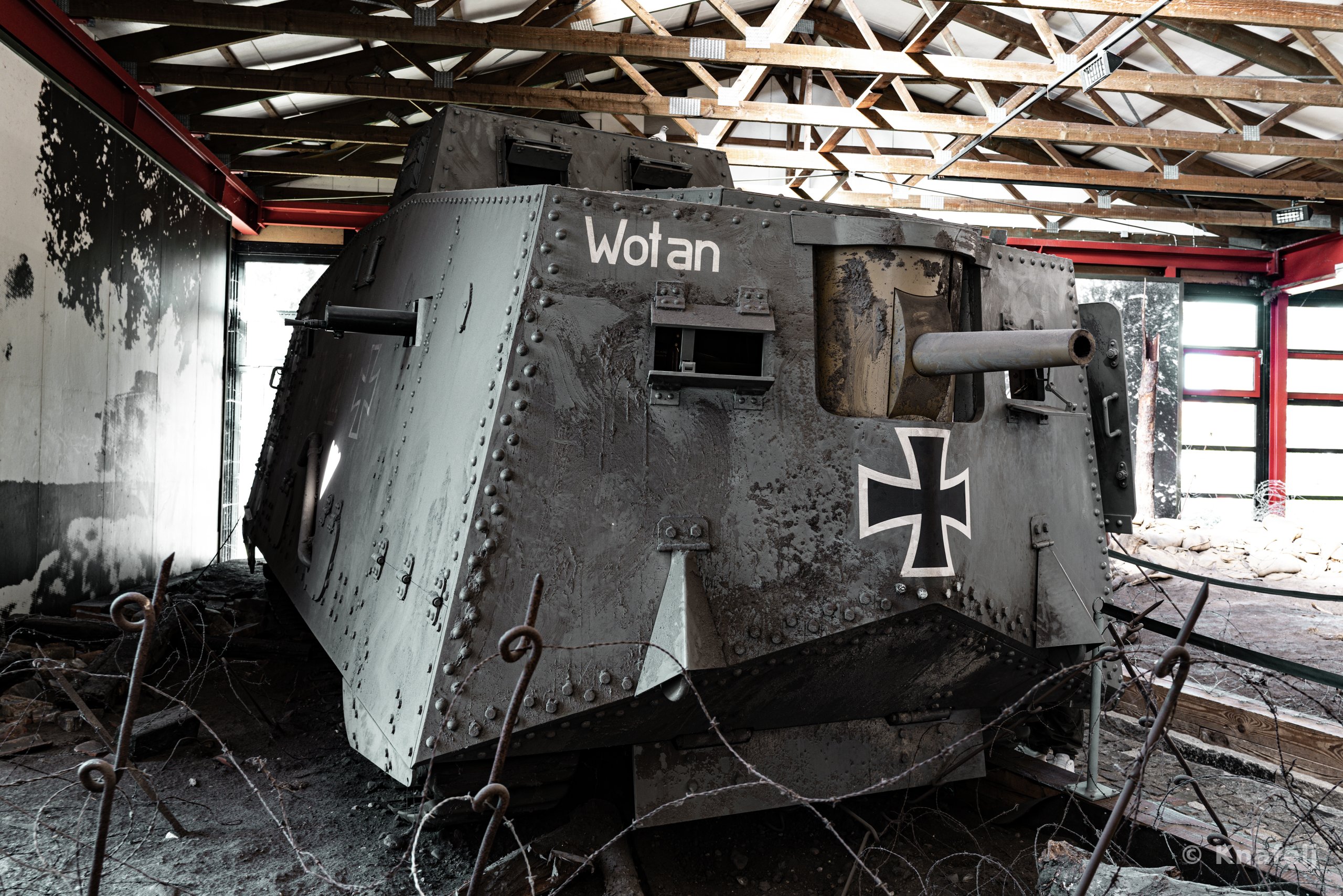 A large grey World War I German tank with the name 'Wotan' and an Iron Cross painted on its side, displayed inside a building with an exposed wooden roof and tangled barbed wire in the foreground.