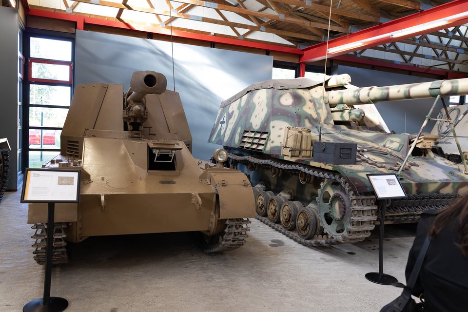 Two World War II-era self-propelled guns displayed indoors in a museum hall with wooden beam ceiling; the left vehicle is painted plain tan and the right one wears green and brown camouflage, each with an informational placard on a stand in front.