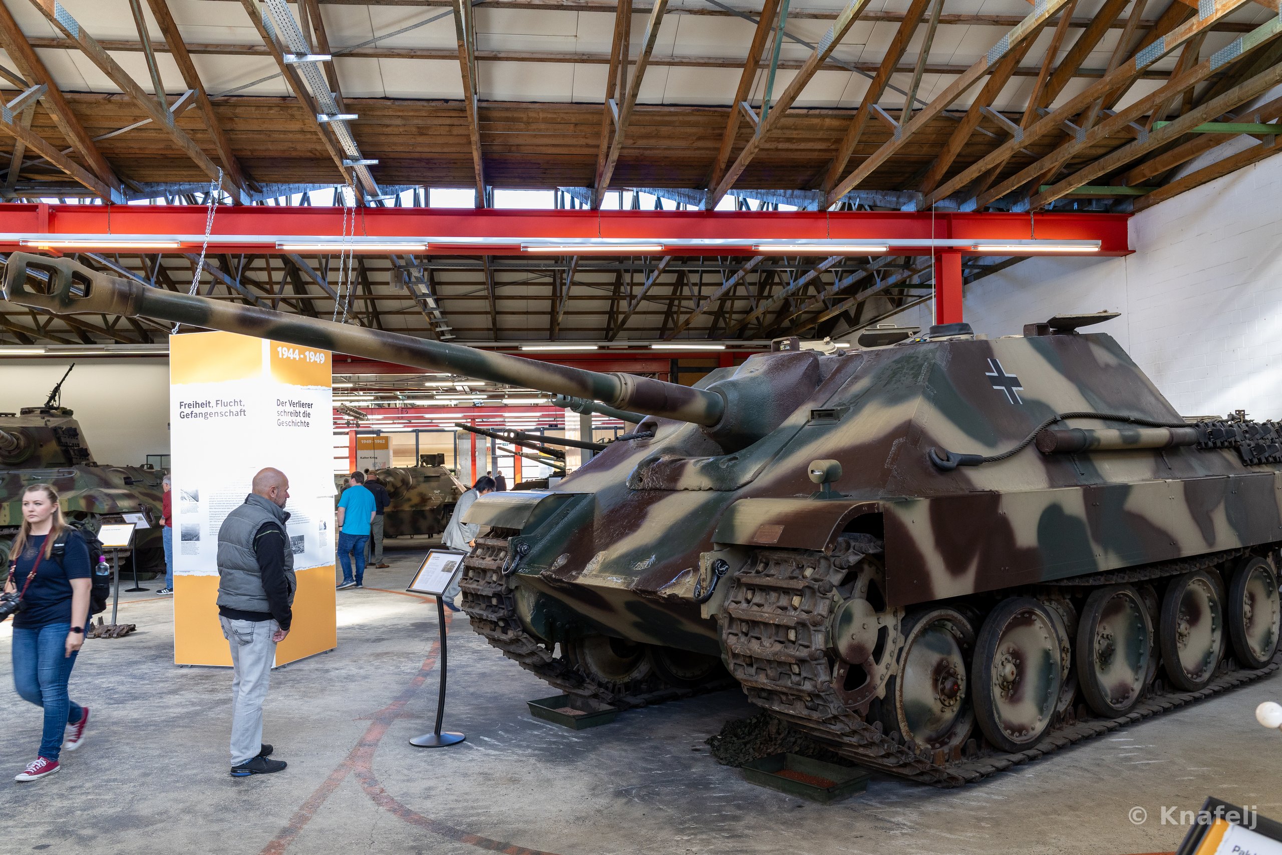 A large camouflaged World War II German tank destroyer with a long gun barrel parked indoors in a museum, with people standing near an orange exhibition sign and other armored vehicles in the background.