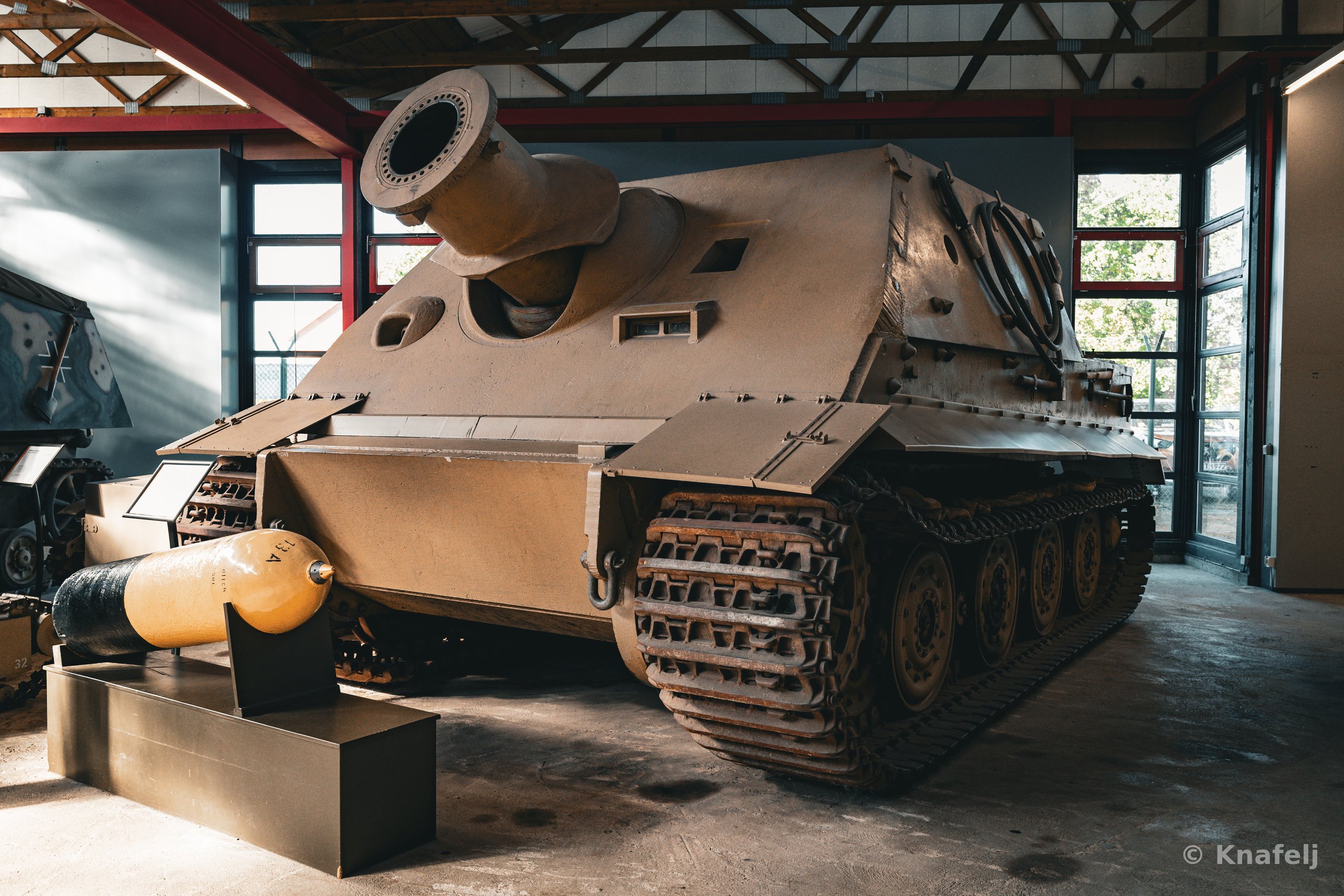 A large heavy armored military vehicle with a short, wide gun barrel displayed indoors on a concrete floor in a museum, with a shell casing in front and windows in the background.