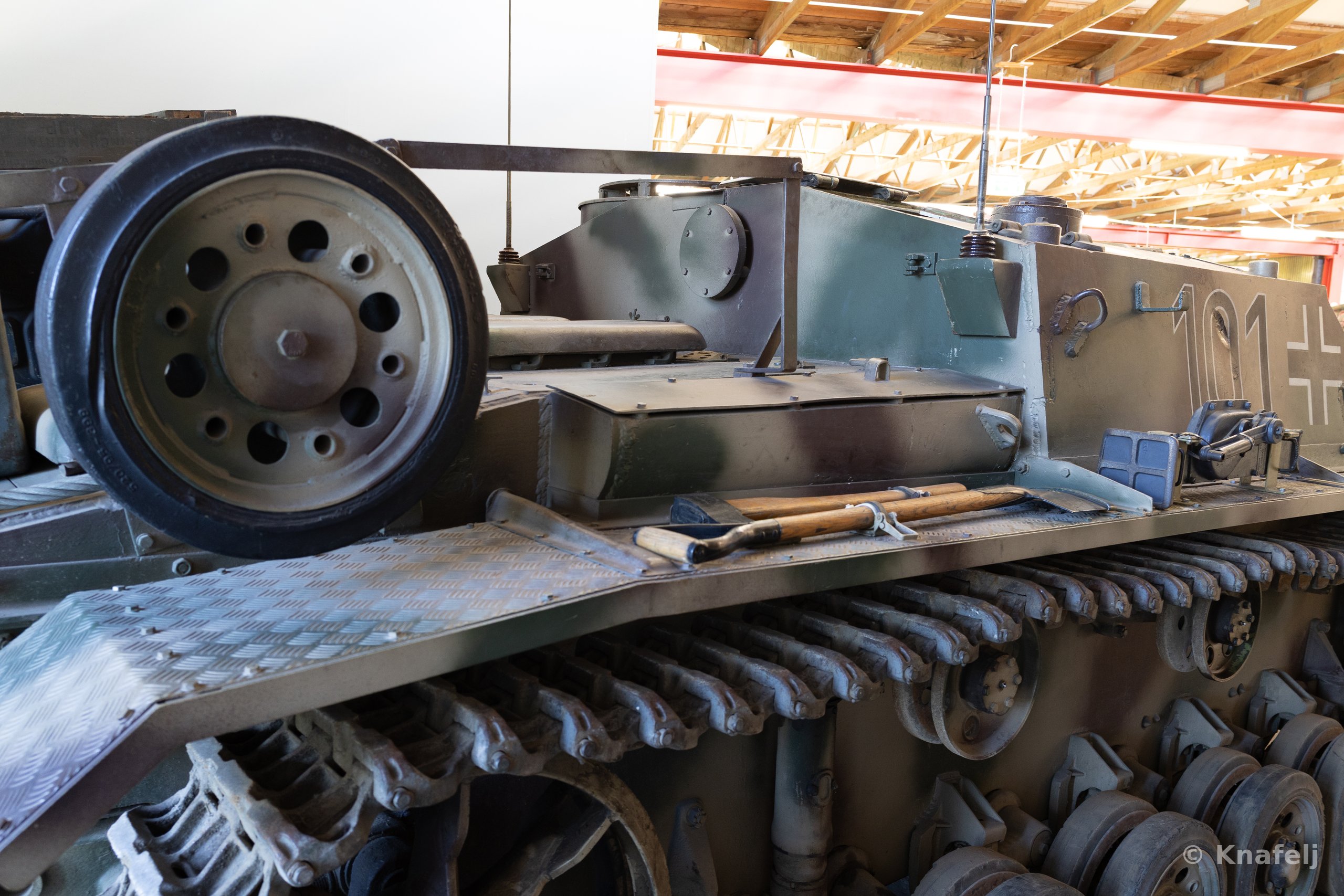 Close-up of a green military tank's side, featuring a large rubber-rimmed road wheel, segmented metal tracks, tools on the fender, and a black-and-white iron cross with the number 101, inside a building with wooden ceiling beams.