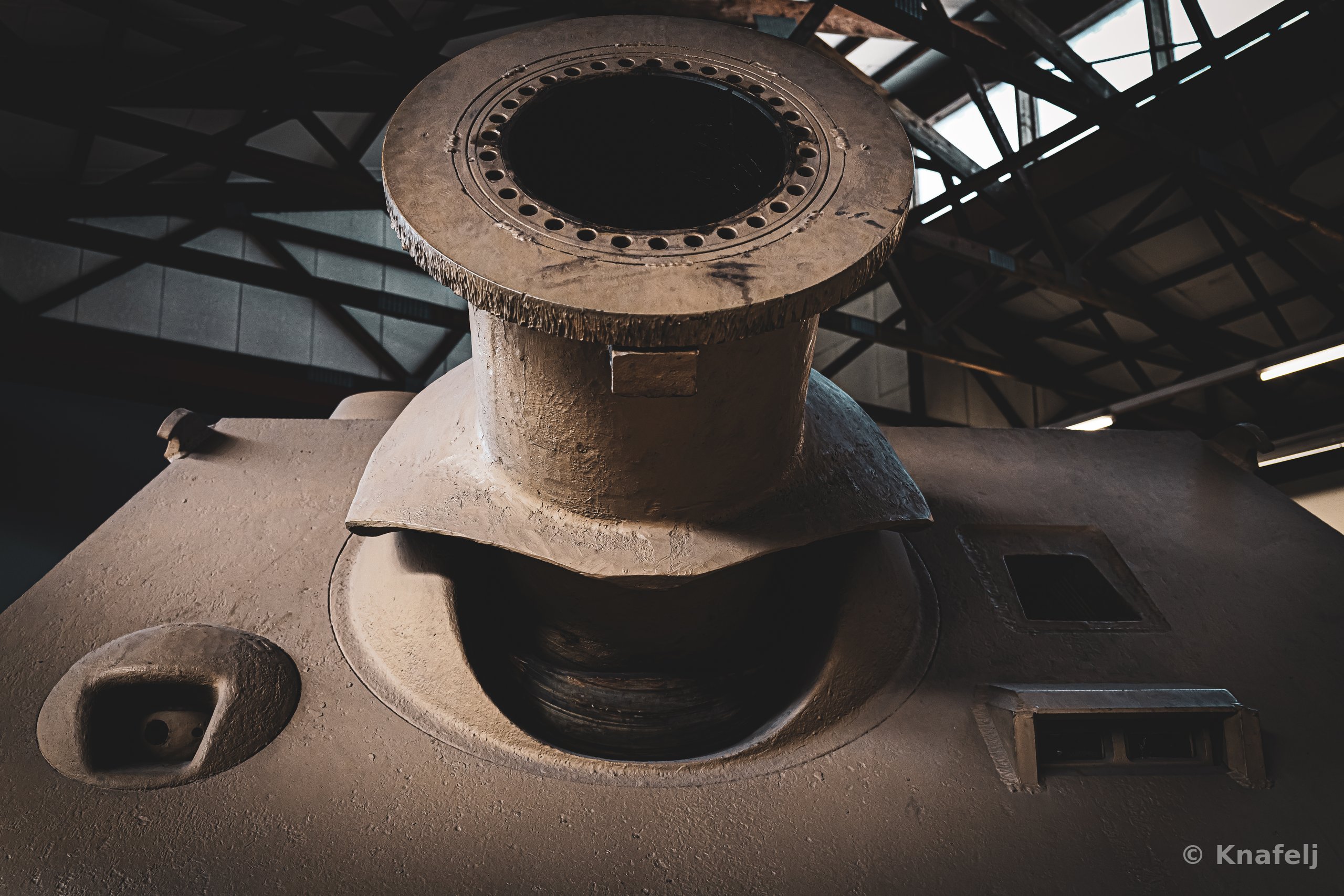 Low-angle view of the large steel turret ring and gun mantlet of a Sturmtiger tank, with bolt holes visible around the rim, set against a dark museum ceiling with metal trusses.