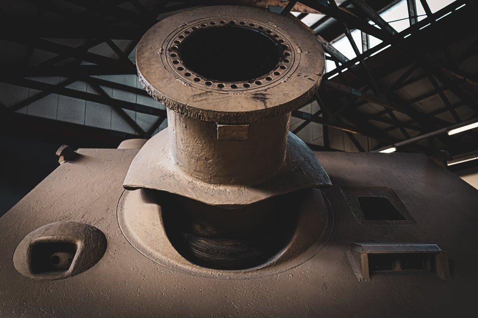 Low-angle view of the large steel turret ring and gun mantlet of a Sturmtiger tank, with bolt holes visible around the rim, set against a dark museum ceiling with metal trusses.