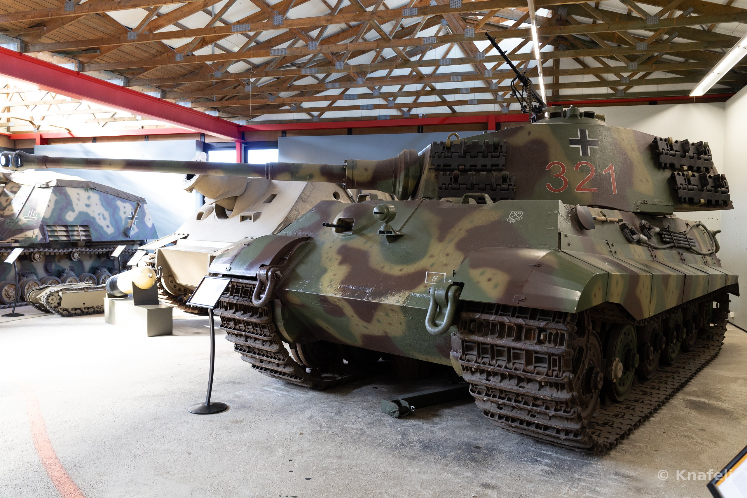 A large World War II German tank with green, brown, and tan camouflage and the number 321 painted in red on its turret, parked indoors in a museum alongside other military vehicles beneath a wooden roof structure.
