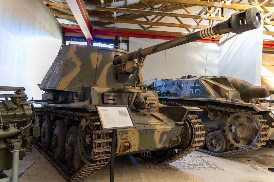A World War II era German Marder III self-propelled gun with a long barrel and camouflage paint, displayed indoors in a museum alongside another armored vehicle marked 101, beneath a wooden-beamed ceiling.