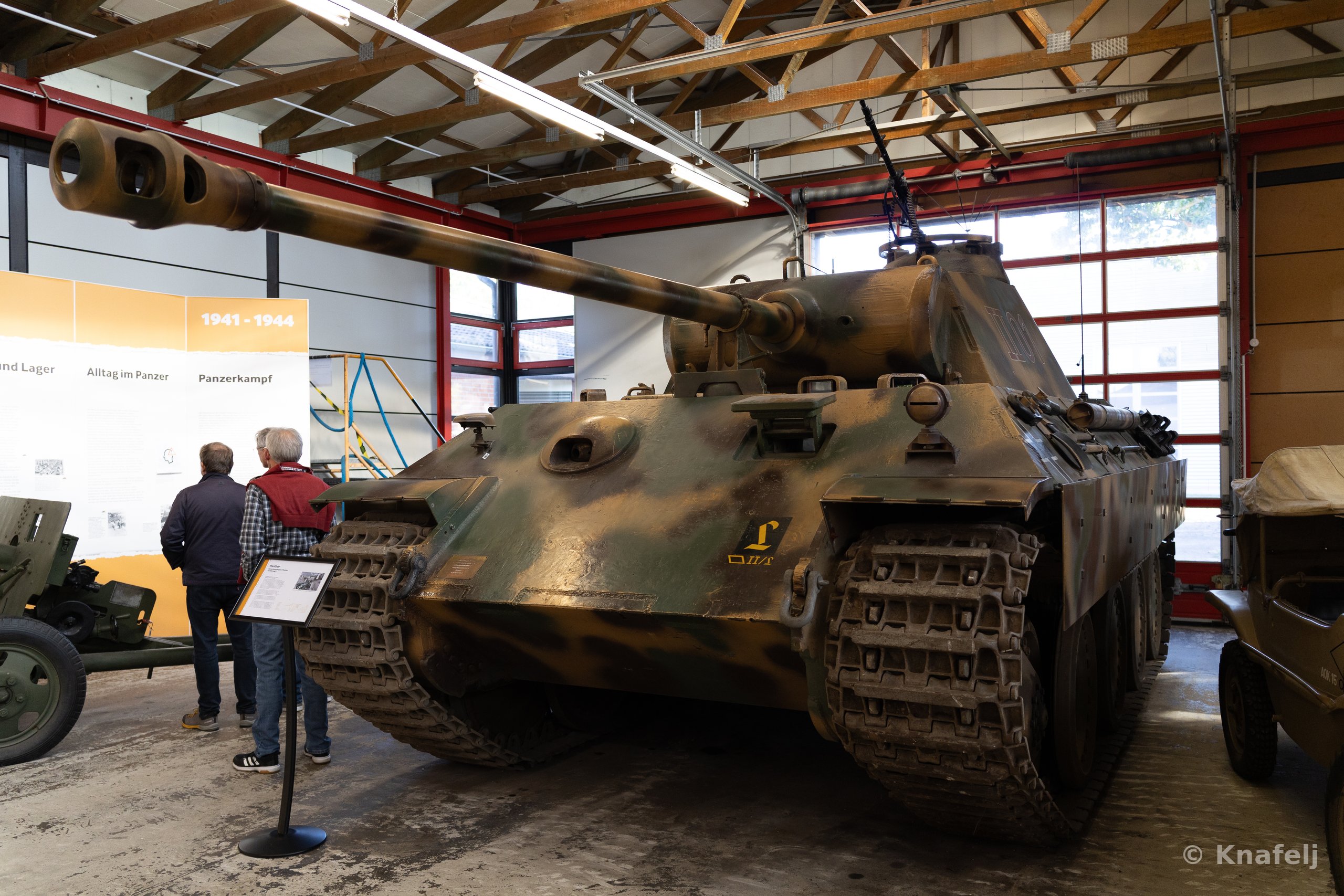 A large camouflage-painted military tank with a long gun barrel displayed indoors in a museum, with two people reading an information sign on the left and exhibit panels on the wall.