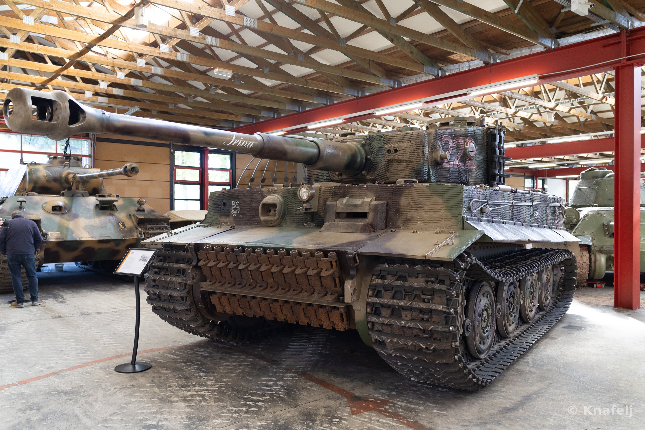 A large camouflaged German Tiger tank with a long gun barrel labeled 'Irina' and the number 222, displayed indoors in a museum with a wooden roof and red support beams, surrounded by other tanks.