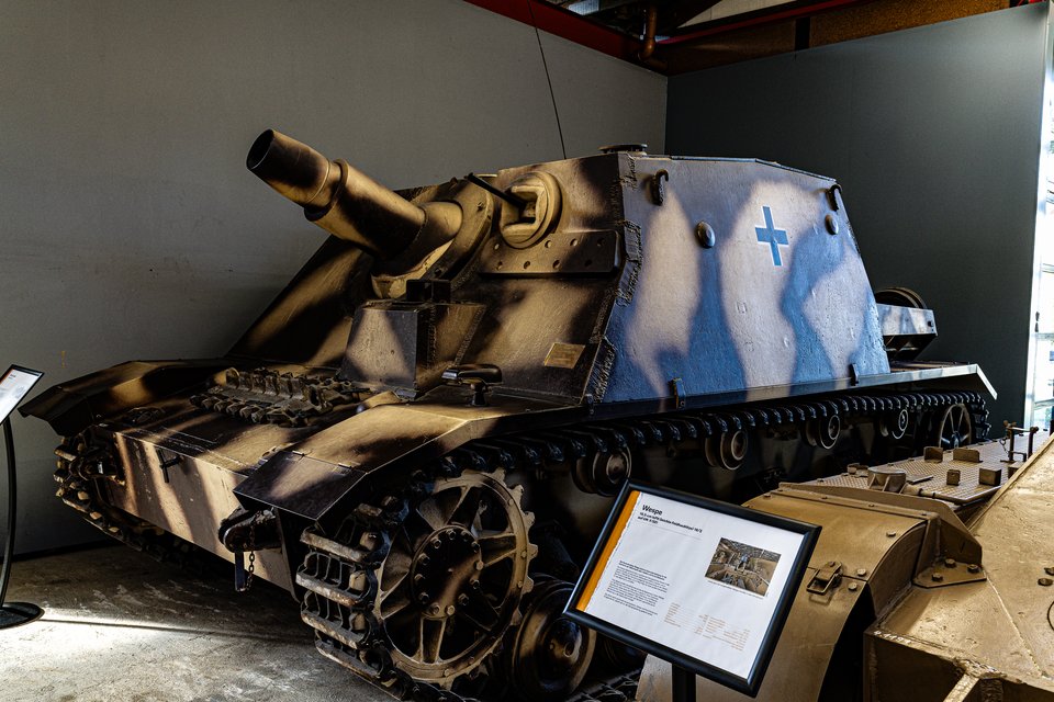 A camouflage-painted tracked military vehicle with a large gun barrel and a blue cross emblem displayed indoors in a museum, with an informational sign in the foreground.