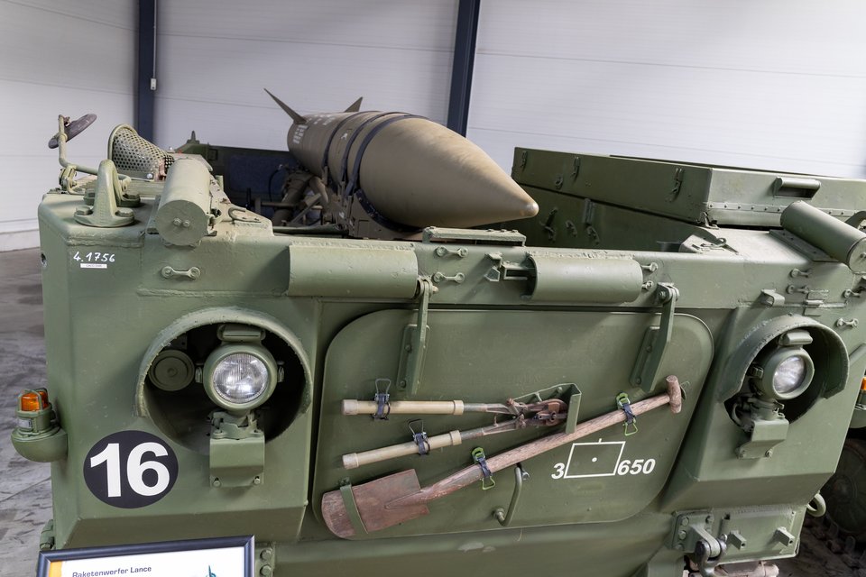 Front section of a green tracked military vehicle bearing a white number 16 on a black circle, with a missile mounted on top, entrenching tools and equipment strapped to the front panel, and round headlights, displayed indoors.