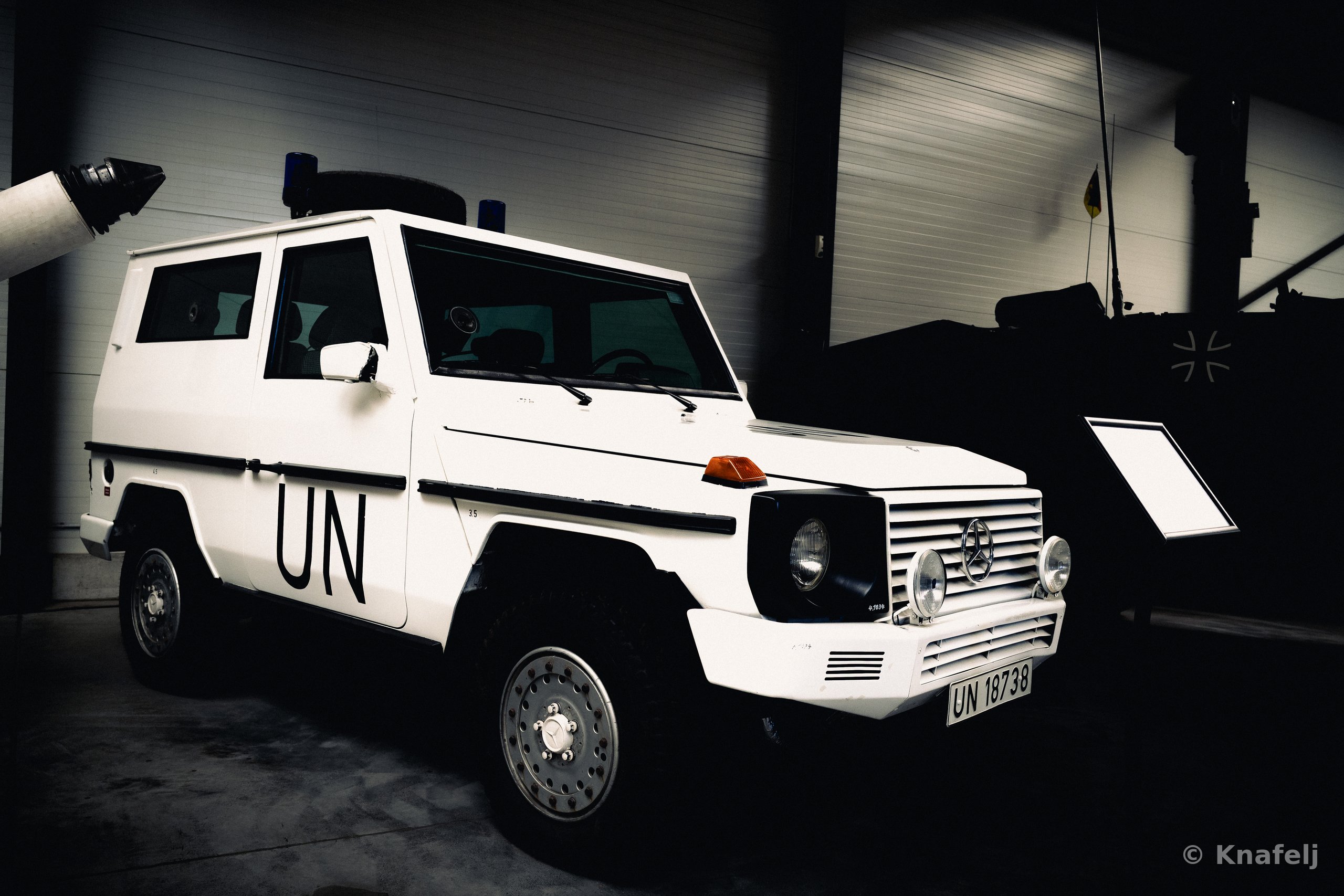 A white Mercedes G-Class military vehicle marked 'UN' on the door with license plate UN 18738, parked indoors in a darkened museum next to a tank and an information placard.