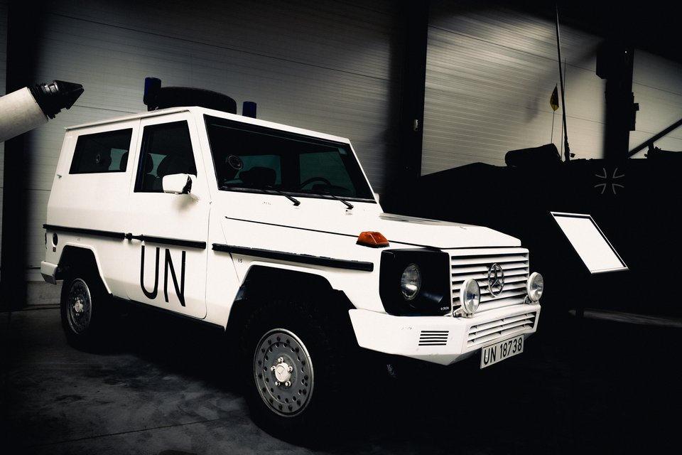 A white Mercedes G-Class military vehicle marked 'UN' on the door with license plate UN 18738, parked indoors in a darkened museum next to a tank and an information placard.