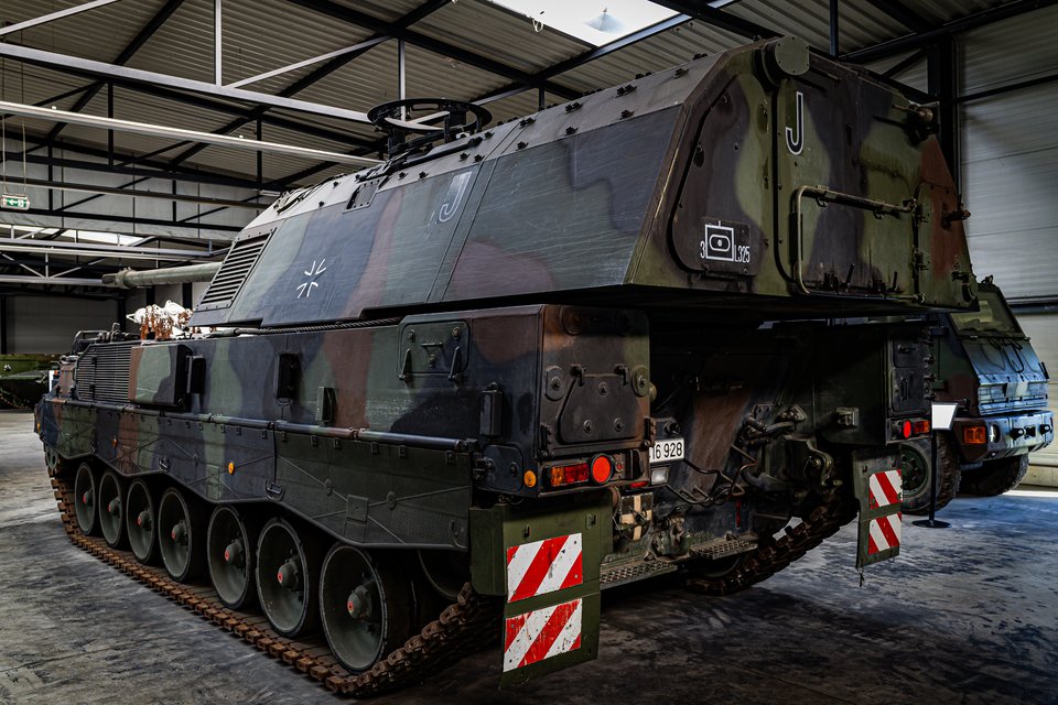 A large green and brown camouflaged tracked military vehicle parked indoors in a museum hall, viewed from the rear, with red and white striped markings and a license plate reading 16 928.