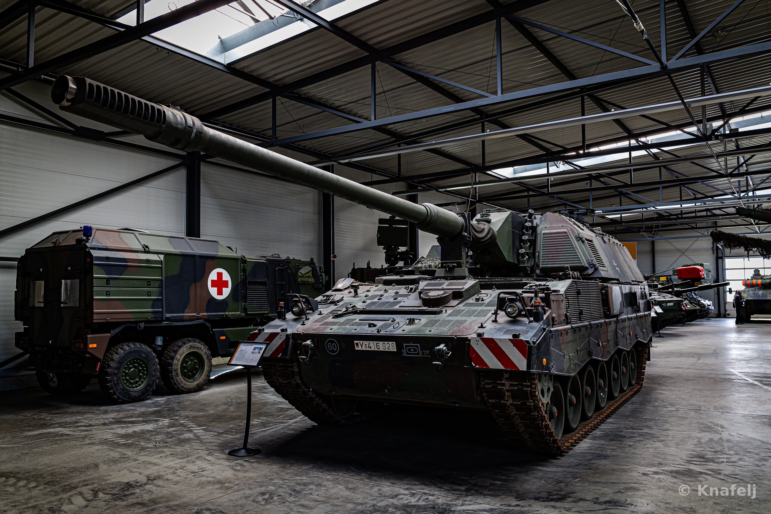 A large green and brown camouflaged self-propelled howitzer with an extended gun barrel parked indoors in a military museum, next to a Red Cross truck and other armored vehicles under a steel-framed roof.