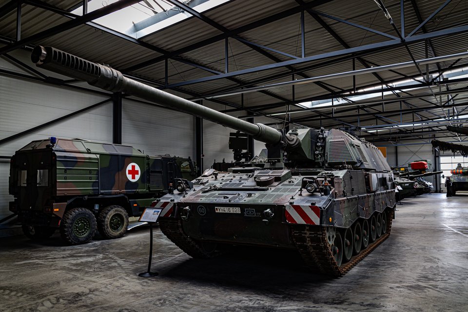 A large green and brown camouflaged self-propelled howitzer with an extended gun barrel parked indoors in a military museum, next to a Red Cross truck and other armored vehicles under a steel-framed roof.