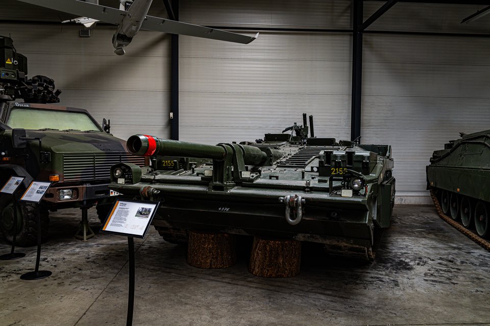 A green military tank with a long cannon and number 2155 displayed indoors in a museum, resting on wooden log supports, with an armored vehicle on the left and informational signage in the foreground.