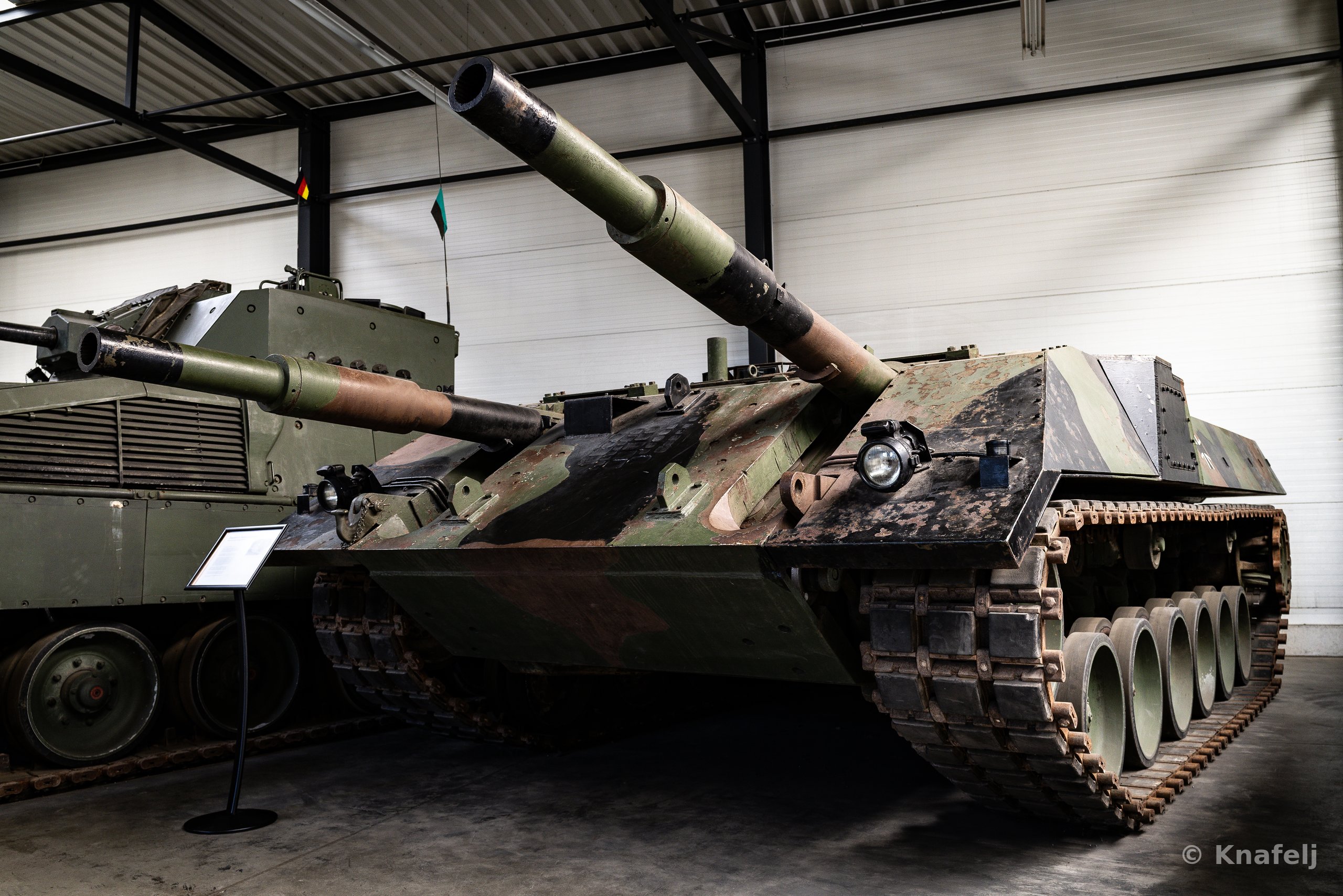 Two military tanks with green and brown camouflage paint parked inside a museum hall, gun barrels pointing forward, with a small informational sign in the foreground.