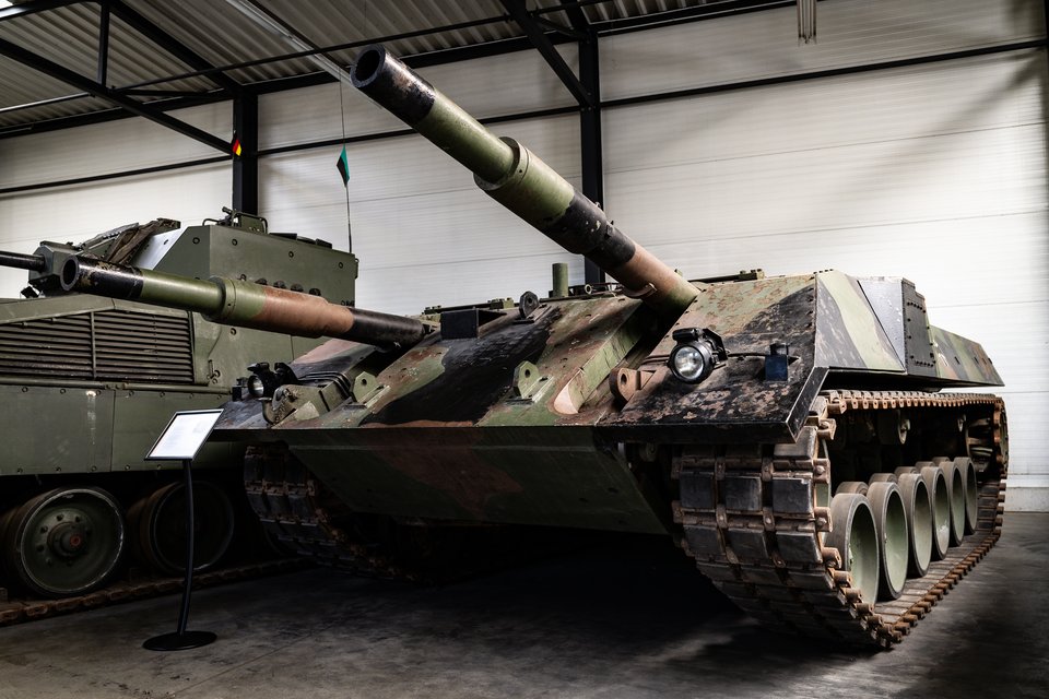 Two military tanks with green and brown camouflage paint parked inside a museum hall, gun barrels pointing forward, with a small informational sign in the foreground.