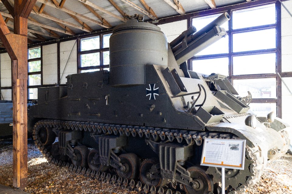 A large gray military tracked vehicle with a long gun barrel and a black Iron Cross insignia, displayed indoors under a wooden roof with windows and an informational sign nearby.