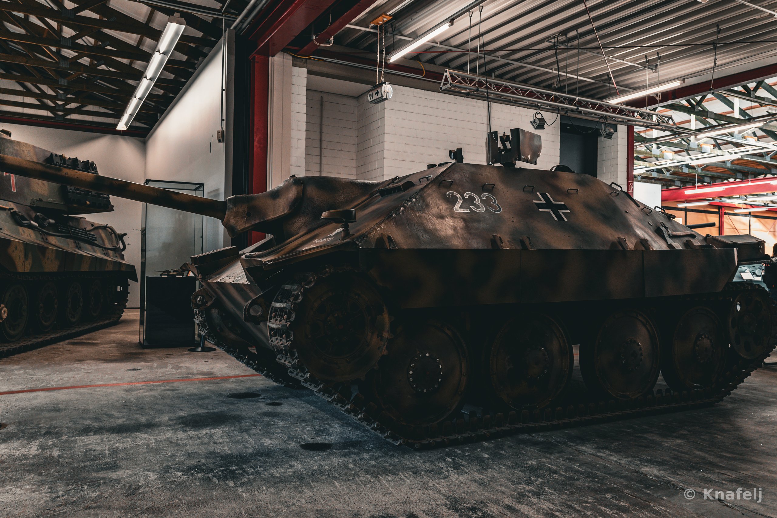 A camouflaged German Hetzer tank destroyer marked with the number 233 and an iron cross, parked indoors in a museum hangar with another armored vehicle visible on the left.