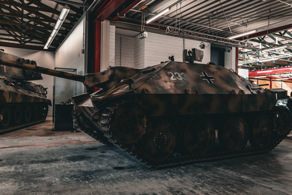 A camouflaged German Hetzer tank destroyer marked with the number 233 and an iron cross, parked indoors in a museum hangar with another armored vehicle visible on the left.