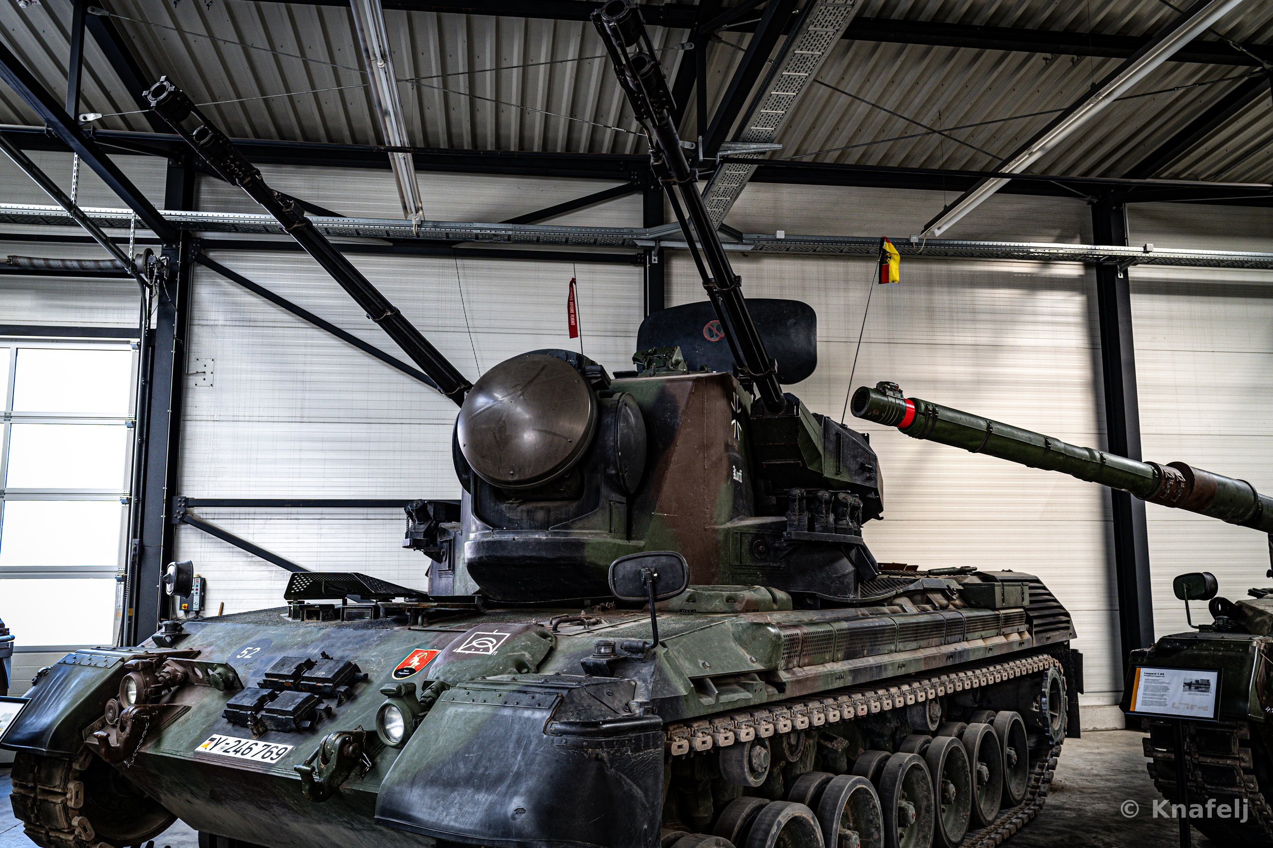 A green and brown camouflaged anti-aircraft tank with twin gun barrels and a rounded radar dome parked indoors in a large hangar with corrugated metal walls and roller doors.