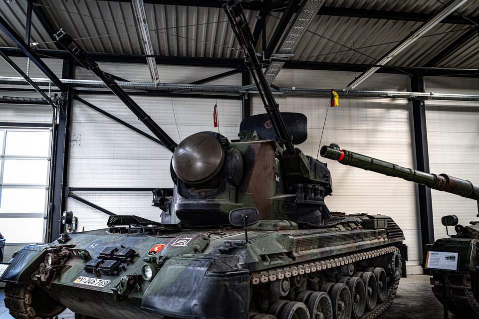 A green and brown camouflaged anti-aircraft tank with twin gun barrels and a rounded radar dome parked indoors in a large hangar with corrugated metal walls and roller doors.