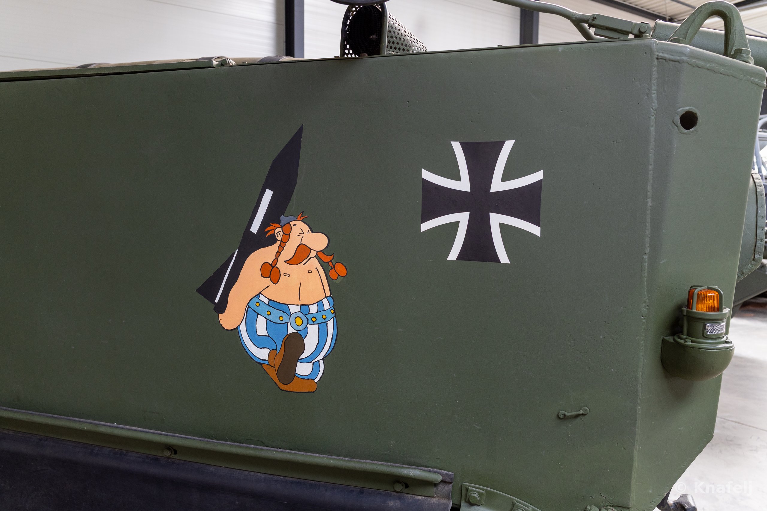 Side panel of an olive-green military armored vehicle featuring a painted cartoon figure with red braided mustache holding a menhir, next to a black and white Iron Cross emblem and an orange marker light.